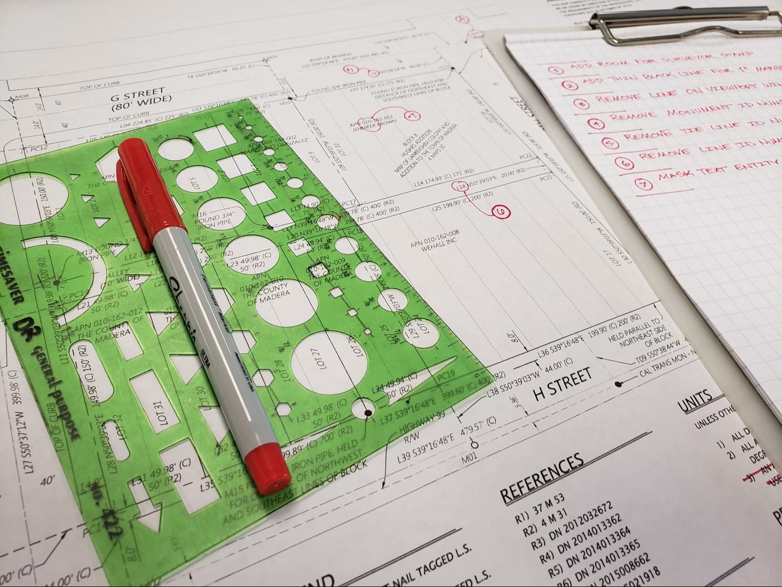 A marked-up drawing set with red-pen redlines and a handwritten review checklist on a clipboard, alongside a drafting circle template.